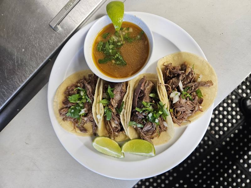 Four birria tacos topped with onion and cilantro, served with a cup of consomé and lime wedges.