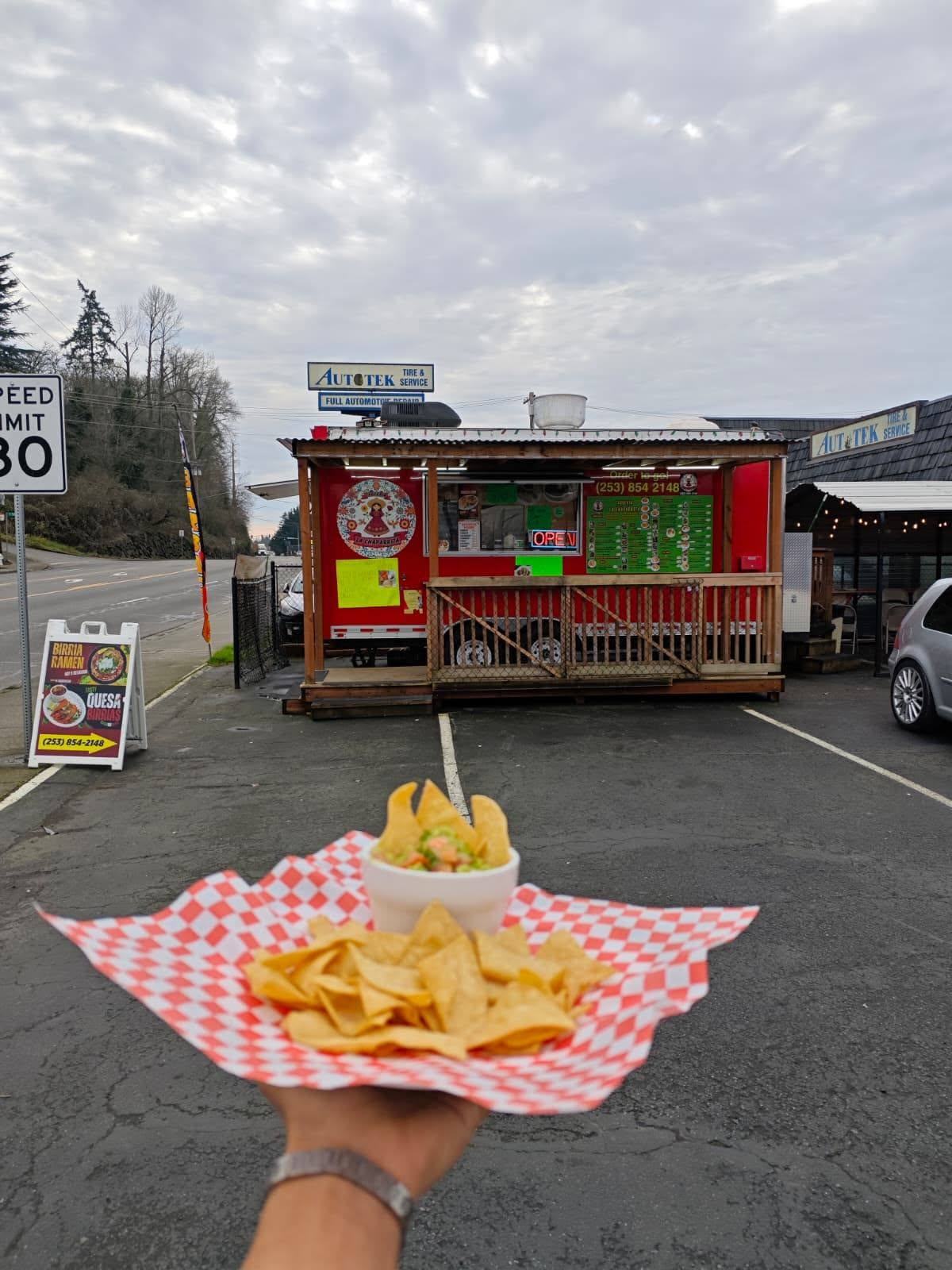 The red La Chaparrita de Kent food truck parked on Central Avenue South, with a basket of tortilla chips and a bowl of dip held up in front of it.
