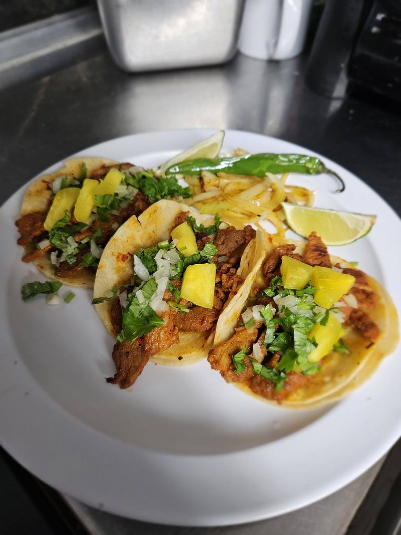 Three tacos al pastor topped with pineapple, onion and cilantro, served with fries, a roasted chile and a wedge of lime.
